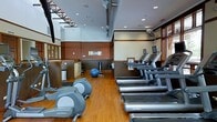 This fitness center offers a thoughtfully designed workout environment that combines functionality with an inviting atmosphere. The space features warm hardwood flooring throughout, complemented by a two-tone wall treatment of rich brown wainscoting and crisp white upper walls that lend a polished, professional feel.

The cardio area is prominently outfitted with a row of Life Fitness treadmills, each equipped with individual display monitors for entertainment and performance tracking. An elliptical cross-trainer provides additional low-impact cardio options. A stability ball and cable resistance equipment round out the functional training zone, catering to a variety of workout styles.

The facility benefits from soaring ceilings with exposed ductwork accents and recessed lighting, creating an open, airy environment. A large overhead fan assists with air circulation during intense workouts. Natural light filters in through slatted wood blinds on the windows, adding warmth to the space. A wall-mounted fan provides additional ventilation. Storage solutions, including a shelving unit, help keep the area organized. The overall design balances a modern fitness aesthetic with warm architectural details, making this a welcoming and fully functional space for daily exercise.