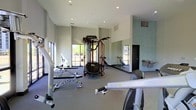 This single-floor fitness center offers a comprehensive workout facility designed to accommodate a range of exercise needs. The space is characterized by soaring ceilings that create an open, airy atmosphere, while large windows along the exterior walls flood the interior with abundant natural light and provide views of the surrounding outdoor environment.

The gym floor is finished with durable, sport-appropriate flooring suited for both equipment use and free movement. A full-length mirrored wall runs along one side of the room, visually expanding the space and allowing users to monitor their form during workouts.

The facility is outfitted with a diverse selection of fitness equipment, including a multi-function cable and strength training machine, adjustable benches, and various resistance-based apparatuses. Cardio equipment is also present, positioned for ease of use throughout the space.

Recessed lighting in the ceiling complements the natural light, ensuring the space remains bright and well-lit at all times. The neutral, light-toned walls and clean layout contribute to a focused and motivating fitness environment. A water fountain is visible near the rear of the space, adding to the functional amenities available to users. Overall, the fitness center is thoughtfully arranged to maximize usability and comfort.