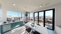 Located at 2395 Herodian Way SE in the Atlanta metro area, this single-level rooftop amenity space combines an interior lounge with a connected outdoor terrace, offering elevated views of the surrounding skyline. The interior is defined by clean, modern finishes, including light tile flooring, recessed lighting, and an abundance of natural light drawn in through floor-to-ceiling glass doors and large windows. A teal-accented wet bar runs along one wall, providing a functional beverage station with ample counter space. A central table with seating accommodates casual gatherings, while framed artwork adds visual character throughout. A dedicated lounge nook features a distinctive mixed-material accent wall composed of brushed metal and marble-style tiles, anchoring a wall-mounted flat-screen television flanked by upholstered seating — an ideal setting for more intimate viewing or conversation. Glass-paneled doors with black steel frames connect the interior seamlessly to the outdoor terrace, which features wood-tile decking, a slatted pergola strung with bistro lights, bar-height seating along the railing, and an outdoor television. The open-air terrace offers unobstructed views of the Atlanta skyline, making it a standout feature of this versatile communal space.