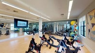 This expansive fitness center is thoughtfully organized across a single floor into distinct workout zones, offering a comprehensive range of equipment and amenities for a full-body training experience. The space features a sleek, modern design with clean white structural columns, high ceilings, and large windows that flood the interior with natural light and provide open views to the outdoors.

The cardio zone is outfitted with a row of stationary cycling bikes, treadmills, elliptical trainers, and rowing machines, all positioned on light hardwood flooring. A large wall-mounted flat-screen TV enhances the cycling and group workout area. Floor-to-ceiling glass partitions elegantly divide sections of the space, maintaining an open, airy feel while creating defined areas.

The strength training section features rubber flooring for safety and durability, and is equipped with a full dumbbell rack, cable machines, weight benches, and selectorized strength equipment. Wall-to-wall mirrors amplify the sense of space and allow exercisers to monitor their form.

A dedicated functional fitness area holds stability balls in multiple sizes, medicine balls, resistance bands, and yoga mats neatly organized on multi-tiered racks. Abstract artwork adorns the walls, adding a touch of personality to the environment. The overall ambiance is bright, professional, and welcoming, catering to a wide range of fitness goals and experience levels.