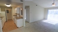 This single-floor residence offers an open and airy layout enhanced by vaulted ceilings that lend a sense of spaciousness throughout. The kitchen is positioned alongside the main living area and features white cabinetry, dark stone-style countertops, a gas range, and a built-in microwave above the stove. The transition between the kitchen and living space is defined by a partial wall divider, preserving an open feel while maintaining a sense of separation. Hardwood-style flooring anchors the kitchen area, while plush carpeting extends through the living room, adding warmth underfoot. Natural light filters in through the windows, brightening the neutral-toned walls and enhancing the overall inviting ambiance. A hallway visible from the main living space leads to additional rooms, including what appears to be a closet with louvered doors — a practical and classic touch. HVAC vents are positioned throughout, ensuring comfortable climate control. The clean, neutral palette of beige and white tones throughout the space provides a versatile backdrop, making it easy to personalize and furnish to a variety of styles.