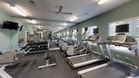 This fitness center is a comprehensively equipped single-floor facility designed to support a wide range of workout routines. The space is divided into distinct functional zones, each thoughtfully arranged to maximize usability. The main cardio area features a row of treadmills and elliptical machines, each equipped with individual monitors, positioned along mirrored walls that visually expand the room. Ceiling fans and fluorescent lighting ensure a comfortable, well-lit environment throughout.

A dedicated cycling studio houses several Peloton-style stationary bikes, each with attached screens, set against full-length mirrors that enhance the sense of space. The sage-green walls and dark carpeted flooring carry consistently throughout, lending a cohesive aesthetic to the facility.

The strength training zone includes a multi-function cable machine, a variety of selectorized weight machines, and a well-stocked dumbbell rack offering a broad range of weights. An open floor area accommodates stretching and functional training, complete with exercise balls and mats visible through a connecting doorway. A water fountain station is conveniently located between zones. Windows along the walls introduce natural light, connecting the interior to the outdoors.