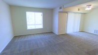 This single-floor space features a open living area finished with wall-to-wall carpeting in a neutral tone, contributing to a clean and understated aesthetic. The room is bathed in natural light through a standard window along one wall, which frames an outdoor view and brightens the interior throughout the day. The walls are painted in a soft, muted green-beige hue, lending a calm and neutral atmosphere throughout. A ceiling-mounted HVAC vent ensures climate control, while the smooth white ceiling keeps the space feeling open. A sliding door fitted with vertical blinds is positioned along one wall, providing access to an adjoining area or exterior space while offering privacy and light control. The layout is straightforward and functional, with clean lines and minimal architectural detailing. Baseboards frame the perimeter of the room, adding a modest finishing touch. Overall, the space presents a simple, practical interior suitable for a range of furnishing arrangements, with its neutral palette and carpeted floors providing a versatile foundation for personalization.