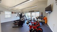 Located at 21 Henry St in Norwich, Connecticut, this dedicated fitness center is a well-appointed, single-floor space designed to support a full range of workout routines. The room features vaulted ceilings with suspended fluorescent lighting that keeps the area bright and energizing throughout the day. Large windows along one wall allow natural light to pour in while offering views of the surrounding outdoor landscape, creating an open and motivating atmosphere.

The space is comprehensively equipped with a diverse selection of cardio machines, including treadmills, elliptical trainers, and a stair climber, providing ample options for aerobic exercise. A row of red cycling bikes offers a dedicated area for spin-style workouts. For strength training, the center includes a free weight station with a dumbbell rack, weight benches, and a medicine ball storage unit. A wall-mounted flat-screen television is positioned for easy viewing during workouts.

The flooring throughout is dark rubber gym matting, designed for durability and safety. Motivational fitness-themed wall decals and framed artwork are displayed throughout the room, reinforcing an energetic and encouraging environment. The layout is thoughtfully arranged to provide clear separation between cardio, cycling, and strength zones, ensuring comfortable use of all equipment simultaneously.