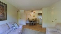 This single-floor residence is designed with an open-concept layout that seamlessly connects the main living spaces. The living area is carpeted and comfortably sized, providing a welcoming space for relaxation. A ceiling fan with an integrated light fixture is centrally positioned, offering both ambient lighting and air circulation throughout the shared space.

The dining area sits adjacent to the living room, featuring wood-look flooring that extends toward the kitchen. It comfortably accommodates a standard dining table and chair set, making it well-suited for everyday meals. Colorful artwork on the walls adds a subtle decorative touch to the space.

The kitchen is efficiently laid out and visible from the dining area, equipped with a white range and overhead cabinetry finished in a warm wood tone. Under-cabinet lighting provides functional task illumination. The textured ceiling runs throughout the unit, adding a classic residential character. Overall, the layout prioritizes functionality and flow, making efficient use of the available space across the interconnected living, dining, and kitchen zones.