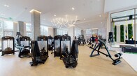 This contemporary fitness center is designed to deliver a premium workout experience within a bright, architecturally refined environment. The main exercise floor is expansive and open, featuring light hardwood-style flooring, soaring ceilings, and floor-to-ceiling windows that flood the space with natural light and provide views of the surrounding urban streetscape. Exposed concrete columns punctuate the floor plan, adding an industrial contrast to the otherwise clean, white interior aesthetic.

The facility is well-equipped with a comprehensive array of professional-grade strength machines, free weights, and resistance equipment arranged throughout the main floor. A dedicated cardio section lines the perimeter windows and includes rowing machines, StairMaster units, and cycling equipment. A functional training area features TRX suspension systems, gymnastic rings, and resistance bands mounted to an overhead rig, with large framed mirrors enhancing visibility and space.

A standout design feature is the dramatic living green wall near the entrance, comprised of vertical moss and fern panels set against a light stone backdrop, creating a striking biophilic focal point. The reception desk is positioned at the entry alongside this installation. A sculptural curved LED chandelier serves as an artistic centerpiece overhead. Floor-to-ceiling glass entry doors with warm wood frames open to an outdoor plaza, seamlessly connecting the interior to the surrounding environment.