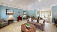 Located at 334 Carolina Forest Blvd in Jacksonville, North Carolina, this single-story community clubhouse offers a well-appointed gathering space designed for comfort and social connection. The interior is characterized by soft teal accent walls, crown molding, and large-format tile flooring that flows throughout the space, creating a cohesive and polished aesthetic.

The main lounge area is a standout feature, boasting vaulted ceilings with recessed lighting and a ceiling fan, a tile-surround fireplace with a classic white mantel, and a wall-mounted television. Natural light pours in through large windows dressed with elegant drape panels, offering views of the surrounding outdoor spaces. Comfortable seating arrangements and a dedicated coffee station make the lounge ideal for casual gatherings.

The full kitchen is equipped with light maple cabinetry, a spacious wraparound countertop, black appliances including a gas range and French door refrigerator, and a generous breakfast bar with counter-height seating — perfect for hosting community events.

Outdoors, the expansive swimming pool is a central amenity, lined with rows of lounge chairs and shaded umbrella seating. A covered pergola structure provides an additional outdoor gathering area. The entire outdoor space is framed by a privacy fence and a backdrop of dense, mature pine trees, offering a serene and private atmosphere.