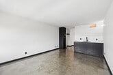 Located in the West Lake neighborhood of Los Angeles, this efficiently designed single-floor unit offers a clean, modern aesthetic throughout. The open living area is characterized by polished concrete floors that reflect natural light, crisp white walls, and dark baseboard trim that adds a sharp contrast to the bright interior. Large windows with roller shades frame views of the surrounding Los Angeles cityscape and greenery, filling the space with abundant natural light.

The living area flows seamlessly into a compact kitchenette, which features slate-blue lower cabinetry, a built-in sink, and a full-height dark refrigerator unit tucked neatly beside an open hallway leading further into the unit. The clean, minimalist design of the kitchen integrates well with the overall aesthetic of the space.

A hallway connects the main living area to the bathroom, which is finished in an all-white palette with small square floor tiles, black matte hardware accents, a wall-mounted towel bar, a toilet, a soaking tub, and a floating vanity topped with a vessel sink beneath a wide mirror. The cohesive use of black fixtures throughout the bathroom and living space ties the unit's design together, creating a contemporary and functional environment well-suited to urban living.