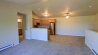 This single-floor space is designed with an open-concept layout that seamlessly connects the kitchen and living area, creating a cohesive and functional environment. The kitchen is equipped with stainless steel appliances, including a full-size refrigerator and a microwave, complemented by warm honey-toned wood cabinetry that extends to the ceiling. A peninsula with a granite-style countertop serves as a natural divider between the kitchen and the living space, offering additional prep and seating opportunities while maintaining the open feel of the floor plan. The kitchen flooring features tile in a neutral earth tone, providing a durable and easy-to-clean surface. Transitioning into the living area, plush wall-to-wall carpet adds warmth and comfort underfoot. A ceiling fan is centrally positioned in the living space, contributing to both airflow and ambient lighting. The walls throughout are finished in a clean, neutral tone, providing a versatile backdrop. Recessed and flush-mount ceiling lights ensure the space is well-lit. The overall ambiance is practical and inviting, suited to everyday living.