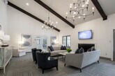 This single-story community clubhouse masterfully blends rustic charm with modern design, creating an inviting and multifunctional gathering space. The architecture is defined by dramatic vaulted ceilings with striking exposed wooden beam trusses, which lend the interior a barn-inspired warmth while maintaining a clean, contemporary aesthetic throughout.

The main gathering area serves as the heart of the space, featuring herringbone-patterned hardwood flooring, a round dining table with upholstered chairs, and a comfortable lounge seating arrangement beneath a stylish chandelier. This open-plan area flows seamlessly into a game room, where a full-size pool table sits beneath high ceilings with ample natural light pouring in through large, dark-wood-framed windows.

A well-appointed kitchenette runs along one side of the space, outfitted with custom gray cabinetry, a wall-mounted television, and a coffee station, making it ideal for hosting events or casual gatherings. Adjacent to this is a dedicated workspace featuring a reclaimed wood accent wall and a built-in desk with a computer workstation.

Generous windows throughout flood the interior with natural light and offer views of the surrounding community outdoor areas. Glass double doors provide direct access to an exterior patio. Warm light wood flooring, white shiplap walls, and rich dark wood accents create a cohesive and welcoming ambiance throughout.