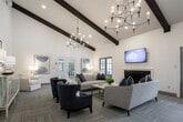 This single-story community clubhouse masterfully blends rustic charm with modern design, creating an inviting and multifunctional gathering space. The architecture is defined by dramatic vaulted ceilings with striking exposed wooden beam trusses, which lend the interior a barn-inspired warmth while maintaining a clean, contemporary aesthetic throughout.

The main gathering area serves as the heart of the space, featuring herringbone-patterned hardwood flooring, a round dining table with upholstered chairs, and a comfortable lounge seating arrangement beneath a stylish chandelier. This open-plan area flows seamlessly into a game room, where a full-size pool table sits beneath high ceilings with ample natural light pouring in through large, dark-wood-framed windows.

A well-appointed kitchenette runs along one side of the space, outfitted with custom gray cabinetry, a wall-mounted television, and a coffee station, making it ideal for hosting events or casual gatherings. Adjacent to this is a dedicated workspace featuring a reclaimed wood accent wall and a built-in desk with a computer workstation.

Generous windows throughout flood the interior with natural light and offer views of the surrounding community outdoor areas. Glass double doors provide direct access to an exterior patio. Warm light wood flooring, white shiplap walls, and rich dark wood accents create a cohesive and welcoming ambiance throughout.