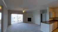 This single-floor living space centers around an open-plan layout that balances comfort with functionality. The main living area is finished with plush wall-to-wall carpeting in a neutral tone, creating a warm and inviting atmosphere. Crown molding runs along the upper perimeter of the room, adding a refined architectural detail that elevates the overall aesthetic.

A built-in fireplace serves as the focal point of the main wall, framed simply with a clean mantle shelf, offering both visual interest and a cozy gathering point. Overhead, a ceiling fan provides both lighting and air circulation throughout the space.

Natural light flows generously into the room through a large sliding glass door fitted with vertical blinds, which opens directly onto a private balcony. The balcony offers views of the surrounding building exterior and outdoor surroundings, extending the living area's connection to the outside.

Adjacent to the living area, a partial view of the kitchen reveals a countertop with warm wood-toned surfaces and what appears to be granite detailing, hinting at a functional and well-appointed cooking space. The neutral color palette throughout ties the entire living area together cohesively.