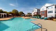 Located at Madison Place Circle in Kernersville, North Carolina, this community amenity space offers residents a well-rounded set of recreational and fitness facilities designed for comfort and active living.

The outdoor pool area is a standout feature, showcasing a generously sized swimming pool with crystal-clear blue water surrounded by a wide concrete deck. Rows of teal lounge chairs invite relaxation under open skies, while two elegant white pergolas provide shaded gathering spots. A children's playground with a colorful slide is visible in the background, and the lush mature trees lining the perimeter create a pleasant, park-like atmosphere. The surrounding multi-story residential buildings, clad in brick and siding, frame the space with a classic, welcoming aesthetic.

The fitness center is thoughtfully equipped to support a variety of workout routines. The main exercise floor features hardwood-style flooring and floor-to-ceiling mirrors that enhance the sense of space. Cardio equipment includes treadmills, ellipticals, and stationary cycling bikes, while a dedicated strength training area offers cable machines, free weights, and benches. Multiple wall-mounted televisions are positioned throughout, allowing residents to stay entertained during workouts. Natural light filters in through slatted blinds, contributing to a bright and energizing environment.