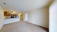 This single-floor residence offers a clean, open-concept layout that maximizes both space and functionality. The living area is finished with neutral-toned wall-to-wall carpeting that creates a warm and inviting atmosphere, while crisp white walls and a smooth ceiling contribute to a bright, airy feel throughout.

The living space flows naturally into the kitchen, which is separated by a practical breakfast bar peninsula — ideal for casual dining or additional counter space. The kitchen is equipped with stainless steel appliances, including a full-size refrigerator and microwave, and features warm honey-toned wood cabinetry that adds a classic, homey character. Pendant lighting above the peninsula provides focused task lighting and a stylish finishing touch.

A doorway from the living area leads to additional rooms, suggesting a well-organized interior layout with clear separation between living and private spaces. The neutral color palette used consistently throughout the unit lends the space a cohesive, move-in-ready aesthetic. Overall, this residence combines practical design with comfortable finishes, making it a functional and welcoming environment suited to a variety of lifestyles.
