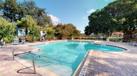 The outdoor pool area is a standout amenity, designed to evoke a resort-like atmosphere. The generously sized rectangular pool features crystal-clear blue water with clearly marked depth indicators, making it suitable for a range of swimmers. The surrounding deck is finished with attractive paver stones, providing a clean and durable surface that complements the overall aesthetic.

Ample seating is available throughout the pool deck, including multiple lounge chairs positioned for sunbathing, along with shaded areas supplied by red market umbrellas — ideal for those seeking relief from direct sunlight. The pool is enclosed by a fence, ensuring safety and privacy, while mature trees lining the perimeter create a natural, lush backdrop that enhances the sense of seclusion.

The open-sky setting allows for abundant natural light throughout the day, contributing to the bright and inviting atmosphere. Surrounding residential buildings are visible in the background, situating the pool as a central community feature. The overall design balances functionality with visual appeal, offering a well-maintained outdoor retreat well-suited for relaxation and recreation. The remaining interior images were too dark to provide additional detail.