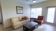 This single-floor space centers around a thoughtfully arranged living room that combines comfort and functionality. The room is finished with smooth, light-colored walls that create a bright and welcoming atmosphere. A ceiling fan with an integrated light fixture is mounted overhead, providing both air circulation and ambient lighting throughout the space.

The living area is furnished with a cushioned sofa in warm beige upholstery, accented with decorative pillows in deep red and patterned tones. A bold burgundy accent chair with white piping adds a contrasting focal point, contributing to the room's layered color palette. A dark-stained rectangular coffee table anchors the seating arrangement, while a matching end table holds a classic table lamp that supplements the natural light.

A large window with white horizontal blinds allows generous natural light to fill the room and overlooks what appears to be an outdoor balcony or railing beyond. A framed piece of wall art with green and neutral tones adds a decorative touch above the sofa. The flooring transitions between carpet and hard surface, contributing to the practical, livable character of the space.