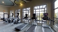 This fitness center is a thoughtfully designed, single-floor facility that offers a comprehensive workout environment for users of all fitness levels. The space features a distinctive ceiling adorned with overlapping circular decorative panels in neutral and muted green tones, adding a modern architectural touch to the room. Recessed lighting throughout ensures the space is well-lit and energizing.

Along one wall, a bank of large grid-pane windows floods the cardio zone with natural light and provides open views of the surrounding outdoor greenery and covered pergola area. This section houses a row of Matrix-brand treadmills, elliptical machines, and stationary bikes, all equipped with individual digital displays.

The strength and functional training area features a full dumbbell rack with a range of weights, a multi-use adjustable workout bench, and a wall-mounted flat-screen television. A large framed mirror reflects the space, enhancing the sense of openness. An organized equipment storage rack holds kettlebells, medicine balls, resistance bands, TRX suspension trainers, and yoga mats, supporting a wide variety of functional fitness routines.

The flooring throughout consists of patterned low-pile carpet tiles, providing comfort and traction. The overall ambiance is clean, modern, and purposefully arranged to accommodate both cardio and strength training workouts.