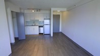 This single-floor unit offers a bright and open-concept layout that seamlessly integrates the kitchen and living areas into one cohesive space. The kitchen is well-appointed with white cabinetry, a mosaic tile backsplash, a stainless steel refrigerator, a dishwasher, and a single-basin sink with a modern faucet. Track lighting above the kitchen provides focused illumination, complementing the recessed lighting near the entryway. The flooring throughout is a warm-toned hardwood-style laminate with dark baseboards, lending a polished and contemporary feel to the space. A textured ceiling adds a subtle design element overhead. The open living area benefits from a generous sense of space, with clean walls and a neutral color palette that creates a calm, versatile atmosphere. The entryway, framed by a structural column, features a solid door and seamlessly transitions into the main living space. The overall design emphasizes functionality and simplicity, making it a practical and inviting environment suited to a variety of lifestyles.