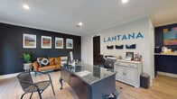 The Lantana office is a thoughtfully designed single-floor professional space that blends bold aesthetics with functional workspaces. The main reception and work area greets visitors with warm light wood flooring, crown molding, and recessed lighting throughout. A striking contrast is created by a deep navy accent wall adorned with a row of framed artwork and a cognac leather sofa, providing a welcoming seating area for guests. A glass-topped executive desk serves as the central workspace, complemented by a white cabinetry unit with gold hardware for organized storage. The bold "LANTANA" branding on the wall reinforces the professional identity of the space, while wall-mounted document organizers and office equipment ensure operational efficiency. A second private office features an additional dark wood executive desk, a wall-mounted display screen, and a door with vibrant teal blue accents that opens to an outdoor view, allowing natural light to fill the room. A colorful abstract painting further energizes this area. The restroom is a standout feature, wrapped floor to ceiling in vivid multicolored brushstroke-patterned wallpaper that adds a lively, artistic touch, paired with a dark vanity and wood-look flooring. Together, these spaces create a cohesive, polished, and personality-driven professional environment.