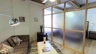 This single-floor space offers a thoughtfully designed interior that seamlessly blends rustic and contemporary elements. Exposed wooden ceiling beams add a sense of character and heritage, while light wood-framed frosted glass partitions create a subtle division of areas, allowing diffused light to filter through and maintain an open, airy feel throughout.

The living area is anchored by light hardwood flooring and features a comfortable seating arrangement centered around a rectangular coffee table, set atop a textured area rug that adds warmth and depth to the space. A dark wood dresser serves as a functional accent piece, complemented by tasteful decorative elements including a tall vase with dried botanicals and a candle holder.

The frosted glass partition walls are a defining architectural feature, providing privacy while preserving the flow of natural light between zones. Overhead, a ceiling fan ensures year-round comfort. The overall palette is neutral and clean, with white walls that allow the natural textures of the exposed beams and wood accents to take center stage. The result is a cozy yet stylish space that balances rustic warmth with a modern, organized sensibility.