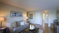 This single-floor space presents a well-appointed living area designed with a neutral and inviting aesthetic. The living room serves as the central gathering space, featuring warm grey walls, a textured ceiling, and hardwood-style flooring that extends throughout the unit, contributing to a cohesive and clean look. A comfortable grey sofa, accented with decorative pillows, anchors the seating area, while a dark leather ottoman doubles as a coffee table, topped with an elegant candle arrangement. Framed black-and-white nautical artwork adorns one wall, adding a classic, curated touch to the space. A table lamp provides warm ambient lighting, complementing the overhead fixtures. A decorative mirror on the adjacent wall enhances the sense of depth in the room. A white paneled door leads to additional areas of the space, with a hallway visible in the background that connects to further rooms, hinting at a practical and straightforward floor plan. The overall ambiance is calm, neutral, and move-in ready, making it a versatile space suitable for a variety of living arrangements.