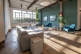Located at 1028 Second Ave S in Birmingham's dynamic Southside district, this single-floor space masterfully blends industrial heritage with contemporary design. The open-plan layout is anchored by soaring ceilings with exposed wooden beams and visible ductwork, lending an authentic warehouse character throughout. Polished concrete floors reflect abundant natural light that pours in through a striking bank of large-pane factory windows along one wall, offering views of the surrounding cityscape and greenery.

The main area is generously proportioned, accommodating distinct zones for seating, dining, and collaborative work. A rustic wood dining table with white metal chairs provides a casual gathering spot, while teal upholstered accent chairs and low acrylic coffee tables define a relaxed lounge area. Lush indoor plants are positioned throughout, softening the industrial aesthetic and adding warmth to the space.

A signature teal accent wall serves as a bold visual centerpiece, complemented by framed black-and-white photography. Vintage industrial shelving units with distressed metal drawers add character and a nod to the building's history. Flexible white curtain dividers allow portions of the space to be sectioned off as needed, offering versatile configuration options. An elevator is also accessible, enhancing the space's functionality.