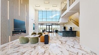Located at 1500 SW Aileron Cir in Bentonville, AR, this contemporary amenity space delivers an impressive first impression through its sweeping, double-height lobby. Polished marble-look tile floors with dramatic veining flow throughout the main area, reflecting abundant natural light that pours in through expansive floor-to-ceiling glass walls and entry doors. The grand lobby features a stylish lounge seating area with rounded velvet chairs and a leather sofa arranged around a coffee table, anchored by a striking wood-paneled accent wall with an integrated fireplace and cascading vertical lighting fixtures. A sleek reception counter clad in teal tile with a stone countertop sits adjacent to the main entrance, and an open staircase with modern railings leads to an upper-level mezzanine. A hallway connects to several additional rooms, including a well-appointed conference room furnished with a long white table, cognac leather rolling chairs, gold pendant lighting, and patterned gold-and-white wallcovering. Glass partitions and frosted panels lend a refined, professional aesthetic to the corridor. A dedicated game room offers a full poker table with padded seating. The overall design balances sophistication with functionality, making it an exceptional shared amenity destination.