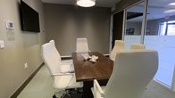 This single-floor conference room offers a refined, professional setting designed for focused meetings and collaborative discussions. The room centers around a rich, dark walnut conference table surrounded by sleek white high-back executive chairs, creating a striking contrast that reinforces a clean, contemporary aesthetic. A cylindrical drum pendant light hangs directly above the table, providing warm, focused illumination that enhances the room's intimate atmosphere. The walls are finished in a deep taupe-gray tone, lending a sophisticated and grounded feel to the space. Neutral-toned carpeting completes the understated palette, contributing to a calm and distraction-free environment. A glass partition wall on one side allows natural light to filter in from an adjacent area, preventing the room from feeling closed off while maintaining privacy. The space is appropriately scaled for small group meetings, accommodating roughly six seated participants comfortably. A decorative white coral accent piece on the table adds a subtle design touch without overwhelming the professional setting. Overall, this conference room balances functional design with polished aesthetics, making it well-suited for client meetings, team briefings, or private discussions.