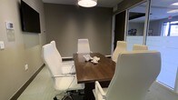 This single-floor conference room offers a refined, professional setting designed for focused meetings and collaborative discussions. The room centers around a rich, dark walnut conference table surrounded by sleek white high-back executive chairs, creating a striking contrast that reinforces a clean, contemporary aesthetic. A cylindrical drum pendant light hangs directly above the table, providing warm, focused illumination that enhances the room's intimate atmosphere. The walls are finished in a deep taupe-gray tone, lending a sophisticated and grounded feel to the space. Neutral-toned carpeting completes the understated palette, contributing to a calm and distraction-free environment. A glass partition wall on one side allows natural light to filter in from an adjacent area, preventing the room from feeling closed off while maintaining privacy. The space is appropriately scaled for small group meetings, accommodating roughly six seated participants comfortably. A decorative white coral accent piece on the table adds a subtle design touch without overwhelming the professional setting. Overall, this conference room balances functional design with polished aesthetics, making it well-suited for client meetings, team briefings, or private discussions.