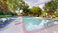 Located at 228 Morrow Rd in Forest Park, Georgia, this outdoor recreational area centers around a generously sized swimming pool with clear turquoise water and classic brick coping along its edges. The pool features multiple entry points via stainless steel handrails, with designated shallow and deep ends clearly marked for safety. Surrounding the pool is a broad concrete deck outfitted with blue lounge chairs and seating, offering ample space for relaxation and sunbathing. A wrought iron security fence encloses the pool area, providing a defined and secure environment. Adjacent to the pool, a well-maintained lawn area provides additional open space, complemented by a shaded patio with umbrella seating ideal for outdoor gatherings. Mature trees — including deciduous and ornamental varieties — frame the entire space, offering natural shade and a lush, park-like atmosphere. A nearby multi-story building with a covered balcony and large glass-panel facade overlooks the pool area, providing convenient access. A children's play structure is also visible within the complex grounds. The overall setting combines functional amenities with green, tree-lined surroundings, creating a welcoming and refreshing outdoor community space.