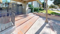 This single-floor unit is part of a well-kept multi-unit residential building characterized by a neutral stucco exterior with contrasting dark shutters and clean architectural lines. The building's exterior is framed by mature trees, including a towering pine, and carefully maintained landscaping featuring ornamental grasses, flowering plants, and a neatly trimmed lawn that creates an inviting, park-like atmosphere.

The entry approach is notable for its classic terracotta brick tile steps, which lead through a covered entryway supported by wooden beam accents. A wrought iron gate provides a sense of security and privacy at the entrance, while additional iron railings line the front steps and perimeter areas, contributing to the building's cohesive aesthetic.

The exterior balconies and windows, framed with dark shutters, add visual depth to the facade and suggest well-proportioned interior spaces with access to natural light. The surrounding streetscape is quiet and suburban, with mature trees visible beyond the property line. Overall, the building presents a tidy, low-maintenance exterior with thoughtful landscaping and classic design details that give it an established, welcoming character.