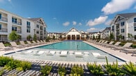 This impressive outdoor amenity space centers around a generously sized resort-style swimming pool with crystal-clear blue water. The pool features built-in in-water lounge platforms and seating, allowing residents to relax directly in the water. A broad, well-finished pool deck surrounds the pool on all sides, lined with rows of modern sling-style chaise lounges and side tables, providing ample space for sunbathing and outdoor relaxation.

At one end of the pool, a striking farmhouse-inspired clubhouse pavilion serves as the architectural focal point, featuring gabled rooflines, large windows, and covered entry areas that open toward the pool. The surrounding multi-story residential buildings echo this modern farmhouse aesthetic, with white board-and-batten siding, dark-trimmed windows, and private balconies that overlook the pool courtyard.

Thoughtful landscaping enhances the ambiance throughout, with ornamental grasses, flowering plants, and young trees planted in decorative gravel beds along the pool's perimeter. A shaded pergola area with umbrella-covered outdoor dining tables provides a comfortable retreat from the sun. The entire space is enclosed by a clean black metal fence, and tall lamp posts add to the welcoming, upscale atmosphere under open skies.