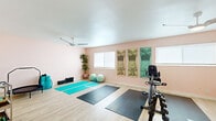 Located in Fremont, CA (94538), this single-floor fitness and wellness space is thoughtfully designed to support a wide range of exercise and wellness activities. The main workout area is generously sized, featuring light hardwood-style flooring that creates a clean, energizing atmosphere throughout. Soft blush-pink walls lend a calming tone to the room, complemented by natural light streaming in through multiple windows.

The workout area is well-organized into distinct functional zones. A dedicated yoga and mat training section is outfitted with vibrant teal yoga mats, foam rollers, and stability balls, while decorative yoga-inspired wall art adds a mindful aesthetic to the space. A ceiling fan ensures comfortable airflow during sessions. On the opposite side of the room, a cardio and strength training zone houses a recumbent exercise bike, a dumbbell rack, and a mini trampoline, all positioned on protective floor mats.

A transitional area features soft pink walls, framed artwork, and a rolling utility cart, serving as a welcoming reception or rest space between the workout zones and connecting rooms. Adjacent to the fitness area, a bright open kitchen with bold orange accent walls, stainless steel appliances, a central island, and sliding glass doors leading to an outdoor area provides convenient access to refreshments. The overall layout is practical, versatile, and conducive to a comprehensive personal wellness routine.