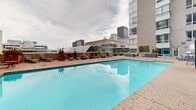 Situated in the heart of downtown Montréal at 1009 Rue De Bleury, this rooftop pool and terrace facility offers a well-appointed urban retreat with impressive views of the city skyline. The centerpiece of the space is a generously sized outdoor swimming pool with crystal-clear blue water, surrounded by a spacious paver-stone deck designed for both relaxation and socializing. Rows of sun loungers line the poolside, providing ample space for guests to unwind while taking in panoramic views of Montréal's iconic buildings and landmarks.

The terrace is organized into distinct zones, each serving a specific purpose. Shaded gazebos with wrought iron dining sets offer comfortable areas for outdoor dining and casual gatherings, while a dedicated BBQ station adds a functional touch to the entertaining space. Lush tropical plants and vibrant floral arrangements are strategically placed throughout, softening the urban surroundings and adding a welcoming, resort-like atmosphere.

A separate lounge area features modular wicker sectional sofas with plush white cushions, arranged around a central fire table — ideal for relaxed conversation or evening gatherings. Cast iron patio furniture tucked into quieter corners of the terrace provides additional intimate seating options. The overall design balances practicality with comfort, making this rooftop terrace a distinctive amenity in the heart of Montréal.