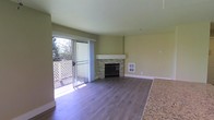 This single-floor space offers a clean, open layout designed for comfortable everyday living. The main living area is a standout, centered around a striking corner fireplace clad in stacked stone with a crisp white mantel surround — a functional and visually appealing focal point that adds warmth and character to the room. Smooth hardwood-style flooring runs throughout, lending a cohesive and modern feel underfoot. Large sliding glass doors flood the living area with generous natural light, while also providing direct access to a private balcony framed by mature trees, creating a serene, nature-connected atmosphere. The balcony features a classic lattice railing, adding a touch of traditional charm. The walls are finished in a soft, neutral tone that complements the natural light and keeps the space feeling bright and airy. A built-in wall vent and subtle architectural detailing reflect thoughtful design throughout. The open sightlines between the living area and adjacent spaces allow for a natural flow, making the layout feel expansive. Overall, this space balances functional design with inviting details, creating a comfortable and well-lit environment.