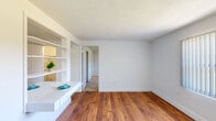 Located at 1410 SW Tenth Terr in Gainesville, Florida, this single-story home offers a clean, functional layout with a consistent design aesthetic throughout. The main living area is anchored by rich hardwood flooring and bright white walls, creating a fresh, open feel. A standout feature is the built-in shelving and desk unit along one wall, providing practical storage and workspace. Natural light filters in through vertical-blind-covered windows, offering outdoor views while maintaining privacy. The entry area also features hardwood floors and a wall-mounted AC unit, keeping the space climate-controlled. The bedroom is finished with wall-to-wall carpet, a ceiling fan with light fixture, and a window with vertical blinds that brings in natural light. The bathroom is a well-appointed space featuring light blue tile wainscoting, wood-look flooring, a vanity with mirror, open built-in shelving, and a shower with curtain — all finished in a clean, cohesive white palette. The kitchen area includes white cabinetry, a double stainless steel sink with granite-look countertops, tile flooring, and a window for natural light. The textured ceilings throughout add a subtle design detail that ties the interior together.