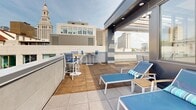 Located atop a contemporary building at 81 Arch St in downtown Hartford, Connecticut, this rooftop terrace offers a well-appointed outdoor retreat with striking views of the city skyline. The space is defined by warm wood decking throughout, complemented by a clean, modern aesthetic featuring white paneled walls and sleek dark metal architectural accents.

The terrace is organized into distinct functional zones. An open-air lounge area provides a generous sectional sofa with white frames and plush cushions accented by bold navy blue pillows, centered around a coffee table — ideal for relaxed gatherings. A wall-mounted flat-screen television adds an entertainment element to this seating nook, which is partially sheltered by an overhead pergola structure with exposed wood beams and a translucent skylight ceiling.

A dedicated sunbathing area features multiple blue chaise lounge chairs arranged to maximize exposure to natural light and panoramic city views. High-top bistro tables and chairs offer an additional casual dining or socializing option in the open-air section of the terrace.

An enclosed glass-walled interior room is accessible via large steel-framed glass doors, providing a climate-controlled counterpart to the outdoor space. Throughout the terrace, sweeping views of Hartford's downtown landmarks — including recognizable historic and modern buildings — serve as a dramatic backdrop, making this rooftop an exceptional urban outdoor amenity.