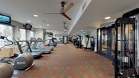 Located at 350 N Ervay St in the heart of Dallas, TX, this well-appointed fitness center offers a spacious, single-floor layout designed to support a full range of workout routines. The main exercise floor is carpeted in a warm geometric pattern and illuminated by recessed lighting and ceiling fans, creating a comfortable and well-ventilated environment for training. A row of treadmills and cardio machines lines one side of the room, while a comprehensive selection of strength training equipment and cable machines occupies the center and opposite wall. Large mirrors along the walls enhance the sense of space and allow for proper form checks during workouts. A dedicated functional training area is set apart from the main floor, outfitted with stability balls, medicine balls, kettlebells, resistance tools, and an elliptical machine. Natural light filters in through multiple windows, offering views of the surrounding Dallas cityscape. A full rack of dumbbells of varying weights is neatly arranged for easy access, accompanied by adjustable benches. The fitness center is accessible through double glass-paned doors, and framed artwork adds a touch of character to the space. The overall design is practical, clean, and well-organized, providing users with everything needed for a thorough and effective workout experience.