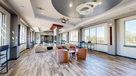 Located at 2525 W Tennessee St in Tallahassee, Florida, this contemporary communal lounge spans a generously sized single floor, thoughtfully designed to encourage both social gathering and relaxed individual use. The open-plan layout is anchored by striking architectural ceiling details, including layered tray ceilings finished in deep gray and white, a sculptural spiral pendant light fixture, and a ceiling fan that adds a functional yet decorative element overhead. Wide-plank wood-look flooring runs throughout, lending warmth and visual continuity to the entire space. Large picture windows line the perimeter walls, flooding the interior with natural light and offering open views of the surrounding Tallahassee landscape. Multiple seating zones are defined within the open floor plan, featuring curved upholstered lounge chairs in warm terracotta tones, modern sofas, and high bar-top workstations positioned along the windows — ideal for focused work or casual conversation. Bold red accent columns and framed artwork punctuate the neutral gray and white walls, adding visual interest and a curated, gallery-like quality. A corridor leads to additional rooms, including a game or activity space visible through a wide architectural opening. The overall ambiance is modern, inviting, and well-appointed, balancing design sophistication with everyday functionality.