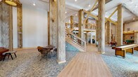 This two-story amenity center showcases a compelling fusion of rustic and contemporary design. The ground floor makes an immediate impression with soaring double-height ceilings, exposed structural timber columns, and bold wooden cross-beam accents that lend the space a warm, barn-inspired character. Light wood-plank flooring and patterned carpet tiles define distinct zones throughout, while large windows flood the interior with abundant natural light and views of the surrounding landscape.

The main level features a generously proportioned open lounge furnished with colorful striped seating, casual round tables, and a striking modular shelving unit displaying curated décor. A dedicated dining area offers comfortable booth-style seating with wall-mounted sconce lighting, ideal for informal gatherings. Adjacent to the lounge, a well-equipped fitness center houses cardio machines, cable weight systems, a dumbbell rack, and strength equipment, all beneath soaring ceilings with ceiling fan ventilation and vivid geometric accent walls.

A wood-and-cable-railed staircase leads to the mezzanine level, which overlooks the main floor below. This upper level provides additional lounge seating, a textured accent wall, and elegant pendant lighting, creating a more intimate setting. Exposed ductwork and industrial-style details complement the natural wood elements throughout, resulting in a cohesive, inviting atmosphere across both floors.