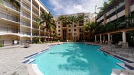 This inviting residential community showcases a striking outdoor amenity space defined by its resort-style atmosphere and tropical surroundings. The centerpiece of the exterior is a generously sized swimming pool with clear, turquoise water, set within a well-maintained courtyard finished with decorative paver decking. Lounge areas and seating are positioned poolside, providing comfortable spaces for relaxation and leisure. Towering royal palm trees line the courtyard, adding lush, tropical character and a sense of established grandeur to the setting. The surrounding multi-story building features warm, earth-toned stucco facades with arched architectural detailing, balconies, and large windows that are characteristic of Mediterranean Revival design — a style well-suited to its sunny Florida setting. A pergola-style structure anchors one side of the pool area, adding architectural interest and shaded outdoor space. The combination of the sparkling pool, verdant palms, and the elegant building facade creates an ambiance that is both refined and relaxed. The overall impression is one of a well-appointed, amenity-rich community designed to offer a high-quality outdoor living experience in a warm-weather environment.