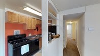 This single-story space offers a practical and well-organized layout centered around a functional kitchen and a carpeted hallway corridor. The kitchen features warm honey-toned wood cabinetry paired with dark granite-style countertops and black appliances, including a gas range. A bold accent wall in a deep terracotta hue adds visual warmth to the cooking area, which is illuminated by overhead fluorescent lighting. A distinctive white built-in shelving unit serves as a partial divider between the kitchen and the hallway, offering open storage space ideal for displaying decorative items or organizing everyday essentials. This architectural detail adds both character and utility to the transitional space. The hallway extends beyond the divider, lined with neutral-toned carpet that continues throughout, contributing to a cohesive and comfortable feel underfoot. Ceiling-mounted light fixtures illuminate the corridor, which leads to additional rooms at the far end. The overall design balances practicality with modest aesthetic touches, making efficient use of the available space while maintaining a functional flow throughout the interior.
