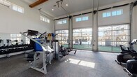 This fitness center is designed to provide a bright, open, and fully equipped workout environment. The space is defined by its dramatically high ceilings, which are accented by exposed wooden beams and clerestory windows that draw in additional natural light from above. A standout architectural feature is the wall of full-panel, floor-to-ceiling glass garage doors that open toward an outdoor pool area, flooding the interior with daylight and offering expansive views of the surrounding community.

The flooring throughout consists of durable, patterned carpet tile suited for high-traffic fitness use. The main workout area is generously proportioned, providing ample room between equipment stations. Cardio equipment is well-represented, with multiple Life Fitness treadmills featuring integrated touchscreen displays, elliptical cross-trainers, and a rowing machine. A dedicated section of the space houses a row of treadmills positioned in front of large windows, enhancing the workout experience with natural views and light.

The strength training area includes a selection of selectorized cable and resistance machines, a full dumbbell rack, weight plates with wall-mounted storage, medicine balls, resistance bands, and a punching bag. Wall-mounted flat-screen televisions are positioned throughout. The overall ambiance is bright, modern, and functional.