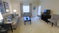 This single-floor space offers a warm and inviting atmosphere highlighted by a thoughtfully designed living area. The living room features soft, neutral-toned carpeting that extends throughout, complemented by light beige walls and classic crown molding that add a refined architectural touch. Natural light filters generously through a large window dressed with elegant floor-length curtains, creating a bright and airy feel. A secondary window with blinds near the entryway door further enhances the room's openness. The living room is centered around a dark wood coffee table that anchors the seating arrangement, which includes a plush sofa and a coordinating loveseat, both in muted gray tones. A patterned accent chair and dark wood media console provide additional function and visual interest. A classic table lamp on a side table adds a soft, warm glow to the space. The overall ambiance is calm and cohesive, with a neutral color palette that creates a sense of comfort and understated elegance throughout. The single-level layout ensures easy accessibility and a seamless flow across the living space.