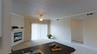 This single-floor residence offers an open-concept design that seamlessly connects the kitchen and living areas, creating a spacious and functional layout. The living room is finished with plush carpeting, neutral-toned walls, and crown molding that adds a subtle architectural detail. A fireplace anchors one wall, providing a focal point and a cozy atmosphere. A large sliding glass door fitted with vertical blinds allows natural light to fill the space and offers potential access to an outdoor area. A ceiling fan with a light fixture provides both ambient lighting and air circulation. The kitchen overlooks the living area and features dark granite-style countertops, a sink, and a kitchen island or peninsula that serves as a natural dividing element between the two spaces. The overall ambiance is clean, bright, and move-in ready, with a simple, understated aesthetic that provides a neutral canvas for personalization. The open sightlines between the kitchen and living room enhance the sense of spaciousness throughout the main living area.