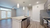 This single-floor residence showcases a contemporary design with a neutral color palette and quality finishes throughout. The open-concept kitchen serves as the centerpiece, anchored by a large island topped with granite countertops, an undermount sink, and pendant lighting overhead. Dark gray cabinetry pairs with a full suite of stainless steel appliances, including a refrigerator, range, microwave, and dishwasher, while white subway tile backsplash adds a clean, polished detail. Wood-look plank flooring extends through the main living areas, complementing the light, airy tones of the walls.

The living area is spacious and well-lit, featuring French doors that invite natural light and provide outdoor access, along with a ceiling fan for added comfort. Multiple pre-wired outlet configurations on the walls offer flexible furniture and entertainment arrangement options.

The bathroom features a wide vanity with granite countertops, navy blue cabinetry, a large mirror, and wood-look flooring that ties cohesively to the rest of the unit. Carpeted bedrooms offer a softer feel, with neutral tones and panel-style doors maintaining the consistent modern aesthetic found throughout the space.