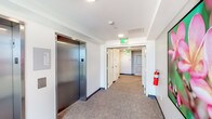 Located at 1221 Ala Kapuna Street in Honolulu, Hawaii, this commercial floor offers a well-appointed and functional layout designed for professional use. The space is accessed via two stainless steel elevators positioned side by side, providing efficient vertical circulation within the building. The elevator lobby opens into a carpeted corridor finished in neutral tones, with clean white walls and recessed lighting that create a bright, polished atmosphere. Dark base molding frames the flooring throughout, lending a refined, cohesive aesthetic to the hallway. Safety features are clearly integrated into the design, including a mounted fire extinguisher and illuminated emergency exit signage. The corridor leads to restroom facilities enclosed by standard commercial-grade doors, completing the functional core of the floor. A standout feature of this floor is a floor-to-ceiling glass window that frames sweeping panoramic views of the surrounding Honolulu landscape, including neighboring commercial rooftops, lush green hillsides, and a glimpse of the coastline beyond. This infusion of natural light and outdoor scenery adds an airy, open quality to what is otherwise a well-structured, professionally designed commercial environment.
