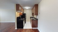 This single-floor space is designed with practicality and comfort in mind. The standout feature is the galley-style kitchen, which makes efficient use of its layout by placing cabinetry, countertops, and appliances along both walls. Rich, warm-toned wood cabinetry lines the upper and lower sections of both sides, providing generous storage throughout. Dark granite-look countertops add a layer of contrast and visual depth to the space.

The kitchen is fully equipped with a black electric range and oven accompanied by an overhead range hood, a stainless steel refrigerator, and a double-basin sink with adjacent lower cabinetry. The flooring transitions from warm hardwood in the adjoining area to clean white tile within the kitchen itself, creating a natural visual boundary between spaces.

Lighting is provided by a central flush-mount ceiling fixture, ensuring the kitchen remains well-lit. The textured ceiling adds a classic touch to the overall aesthetic. Neutral wall tones complement the cabinetry and keep the space feeling bright and open. The layout is highly functional, making the most of the available space while maintaining a clean, organized appearance throughout.