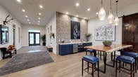 The Marq's single-floor clubhouse showcases a refined modern farmhouse design that seamlessly integrates rustic and contemporary elements. Throughout the space, white shiplap walls contrast with warm wood-look flooring, creating a cohesive and inviting aesthetic. The entry area features a welcoming foyer with large glass double doors, area rugs, and a built-in wet bar station highlighted by navy blue cabinetry, stainless steel appliances, and a striking exposed brick accent wall adorned with equestrian-themed artwork.

The main lounge area is generously proportioned, bathed in natural light from large windows dressed with floor-length draping curtains that offer views of the surrounding property. Seating includes patterned sofas, leather armchairs, and cowhide rugs anchored beneath gold wire-frame chandeliers, lending an upscale yet relaxed atmosphere. Rustic barn doors on sliding hardware connect the lounge to a separate flex room, ideal for private gatherings or work sessions.

A dedicated office or conference area with glass-paneled walls and a wood-framed door provides a functional workspace within the clubhouse. Thoughtfully curated artwork, lush greenery, and layered lighting — including pendant fixtures and recessed lighting — complete the polished, detail-oriented design of this community gathering space.