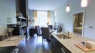 This single-floor residence offers a seamlessly connected open-concept layout that integrates the kitchen, dining, and living areas into one cohesive, well-appointed space. The kitchen is outfitted with rich dark cabinetry, granite countertops, a stainless steel sink, and a sleek pendant light that adds a warm, focused glow over the workspace. Dark hardwood flooring flows continuously throughout, grounding the space with a sense of warmth and sophistication. The dining area features an elegant table surrounded by tall, upholstered chairs in a deep charcoal tone, creating a refined setting for entertaining. An open shelving unit along one wall displays glassware and decorative accents, adding both function and visual interest. The living area beyond is bathed in natural light streaming through large windows dressed with sheer roller shades and floor-length gold-toned drapes, lending an airy, inviting ambiance. The walls are finished in a soft, neutral gray tone that complements the darker furnishings and flooring throughout. Together, the thoughtfully curated design elements create a modern yet comfortable atmosphere that balances style with everyday livability.